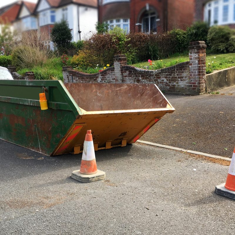 Big rusty industrial skip with plastic safety cones on side of road. Focus on front of the metal bin with space to add text on surrounding area, footpath, road surface & blurry houses in background.